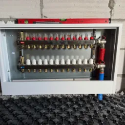 Rozdzielacz ogrzewania podłogowego Ferro w białej szafce, zamontowany na ścianie z bloczk&oacute;w betonowych, z widoczną matą systemową na podłodze.