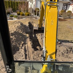 Przemysław Rosół - Żółta koparka z kabiny operatora, widok na wykopany wąski rów w ziemi na tle domu i zieleni. Ślady gąsienic w wykopie. Słoneczny dzień.