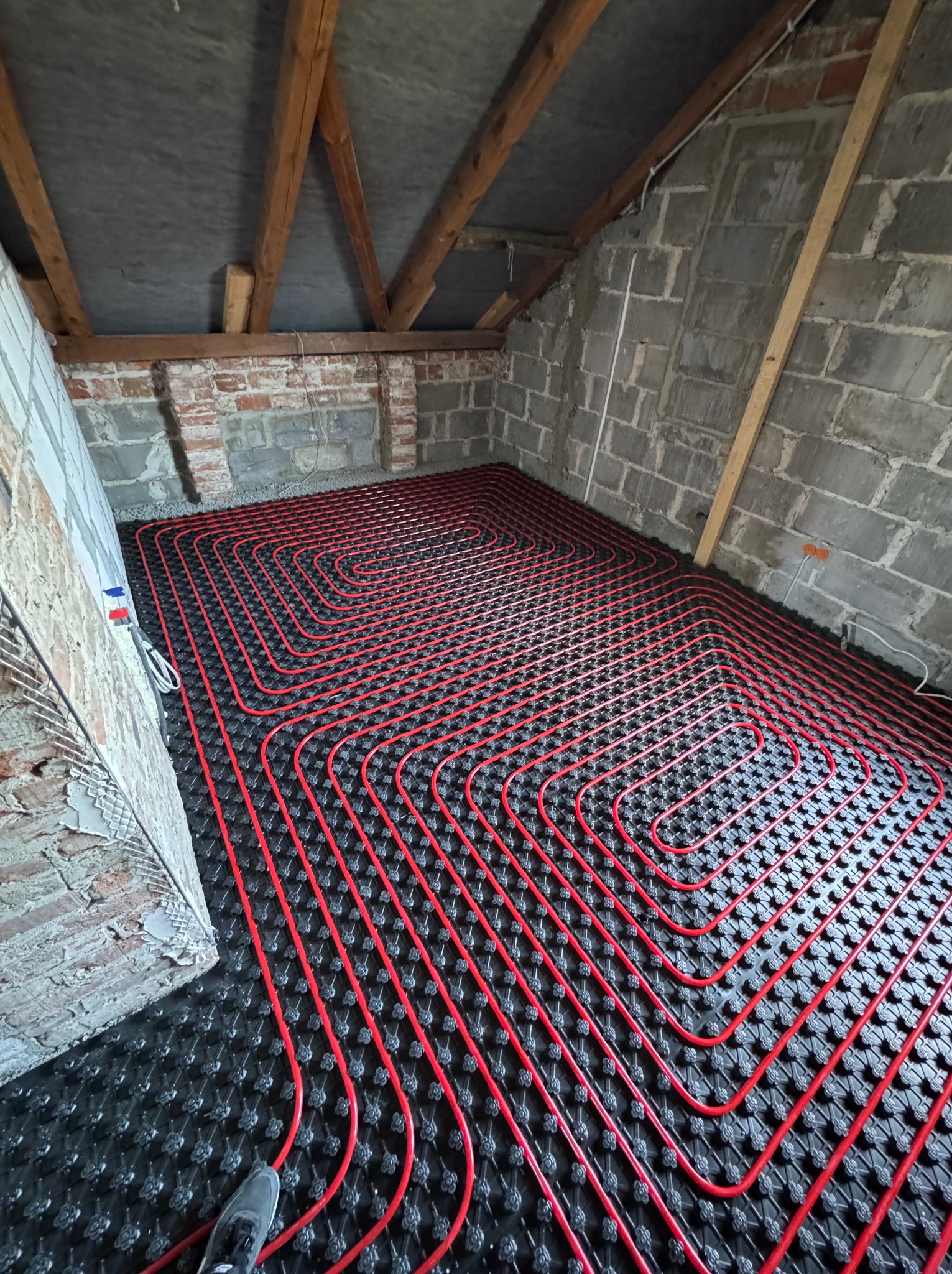 Czerwone rury ogrzewania podłogowego ułożone na czarnej macie w surowym pomieszczeniu z ceglanymi ścianami i drewnianą konstrukcją dachu. Widok z góry na instalację.