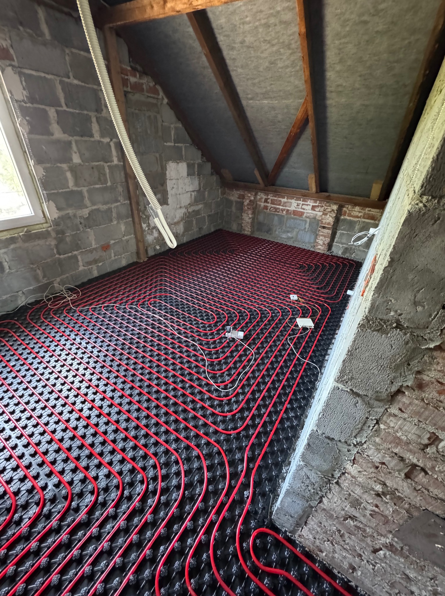 Czerwone rury ogrzewania podłogowego na czarnej macie w surowym wnętrzu poddasza z ceglanymi ścianami i drewnianą konstrukcją dachu. Widoczne przewody i okno.