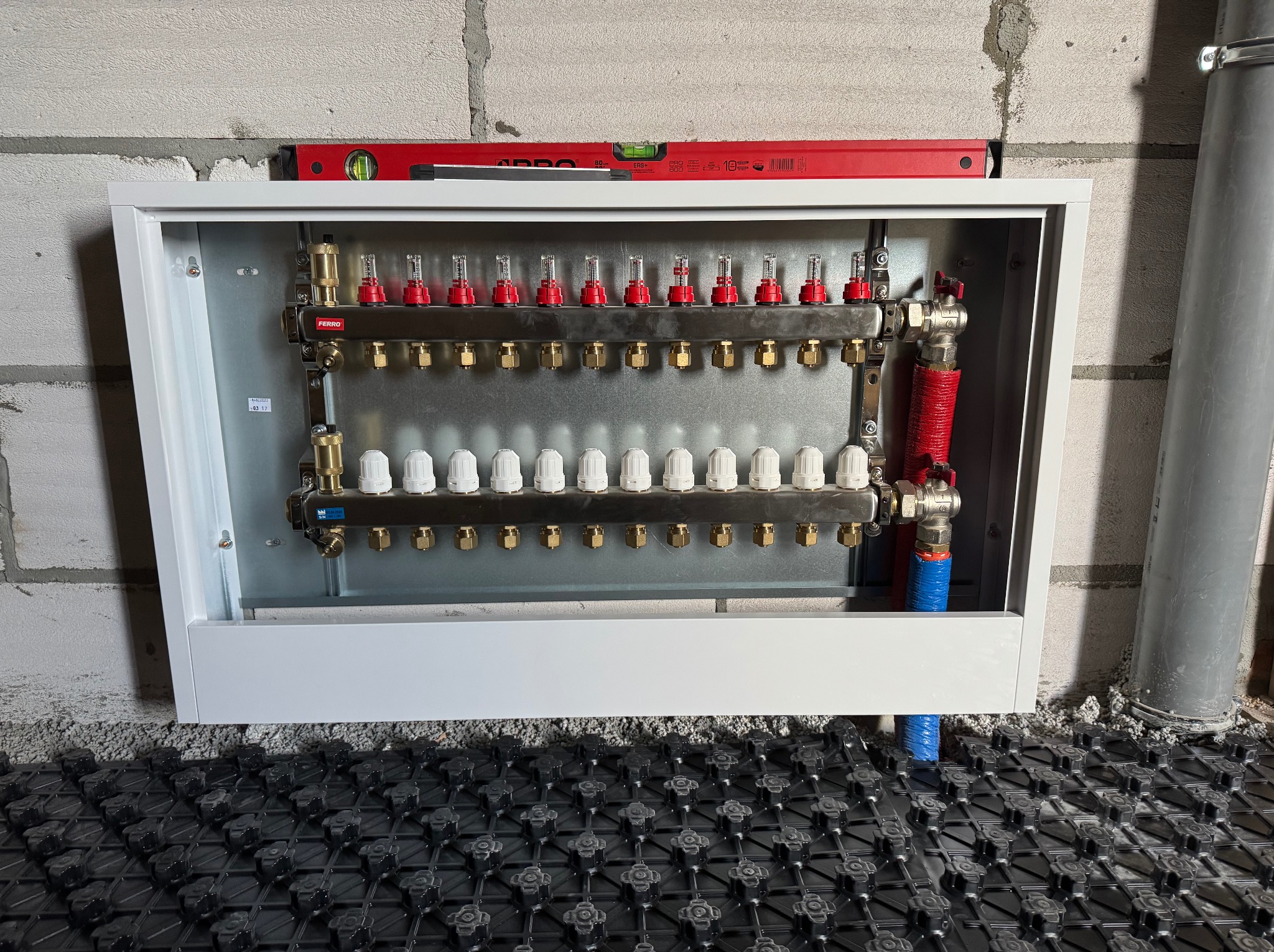 Rozdzielacz ogrzewania podłogowego Ferro w białej szafce, zamontowany na ścianie z bloczków betonowych, z widoczną matą systemową na podłodze.