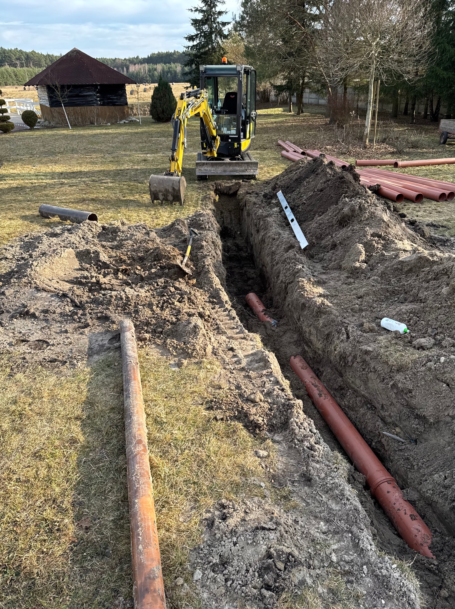 Wykop pod instalację z ułożonymi rurami kanalizacyjnymi w ziemi. Żółta koparka stoi w tle na trawniku. Widoczny poziomica i łopata.