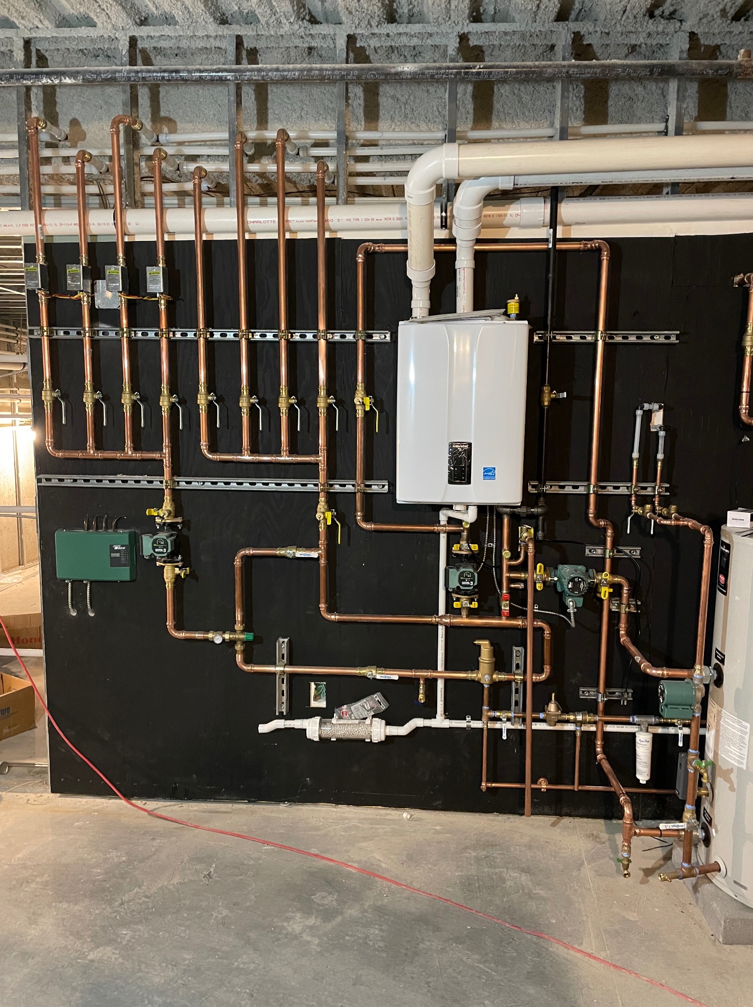 Kompleksowa instalacja hydrauliczna z miedzianymi rurami, zaworami i białym bojlerem na czarnym tle. Widoczne elementy mocujące i fragment surowego sufitu.