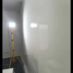 Wnętrze po pracach wykończeniowych. Ściana o jednolitej, gładkiej powierzchni, oświetlona lampą budowlaną. Na podłodze folia ochronna. Widoczny fragment drabiny.