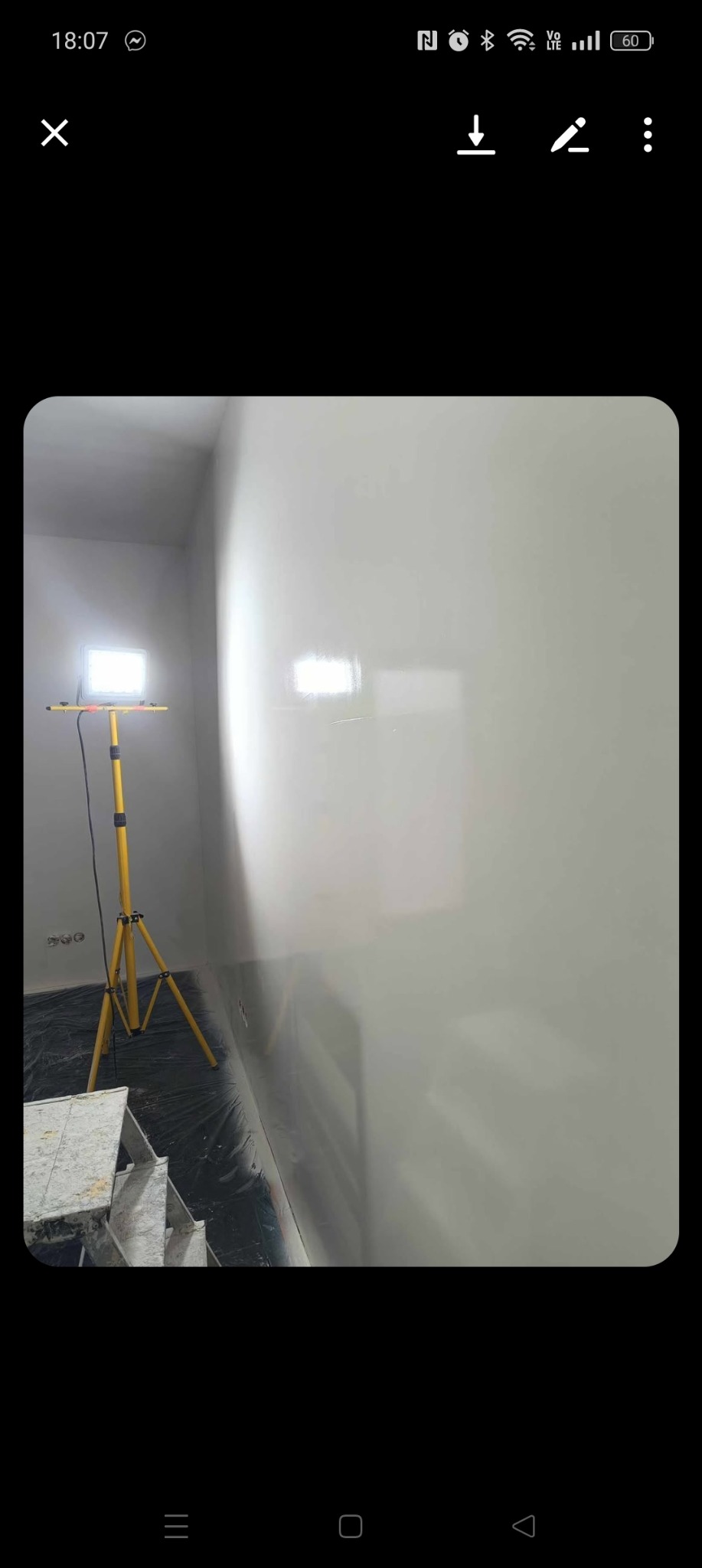 Wnętrze po pracach wykończeniowych. Ściana o jednolitej, gładkiej powierzchni, oświetlona lampą budowlaną. Na podłodze folia ochronna. Widoczny fragment drabiny.