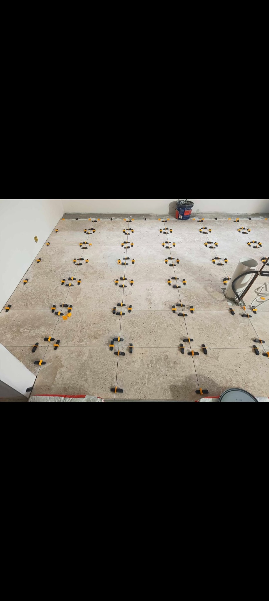 Układanie płytek podłogowych z użyciem poziomujących klipsów i klinów. Widoczny kubeł z zaprawą i narzędzia glazurnicze. Jasne, beżowe płytki w trakcie montażu.