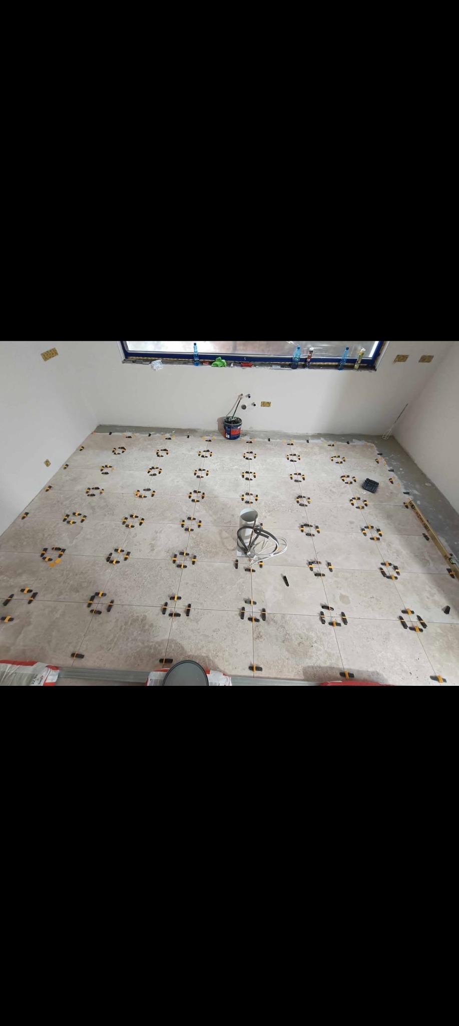 Jasne płytki podłogowe w trakcie układania z użyciem systemu poziomowania, widoczne narzędzia i akcesoria, okno w tle. Prace wykończeniowe w pomieszczeniu.