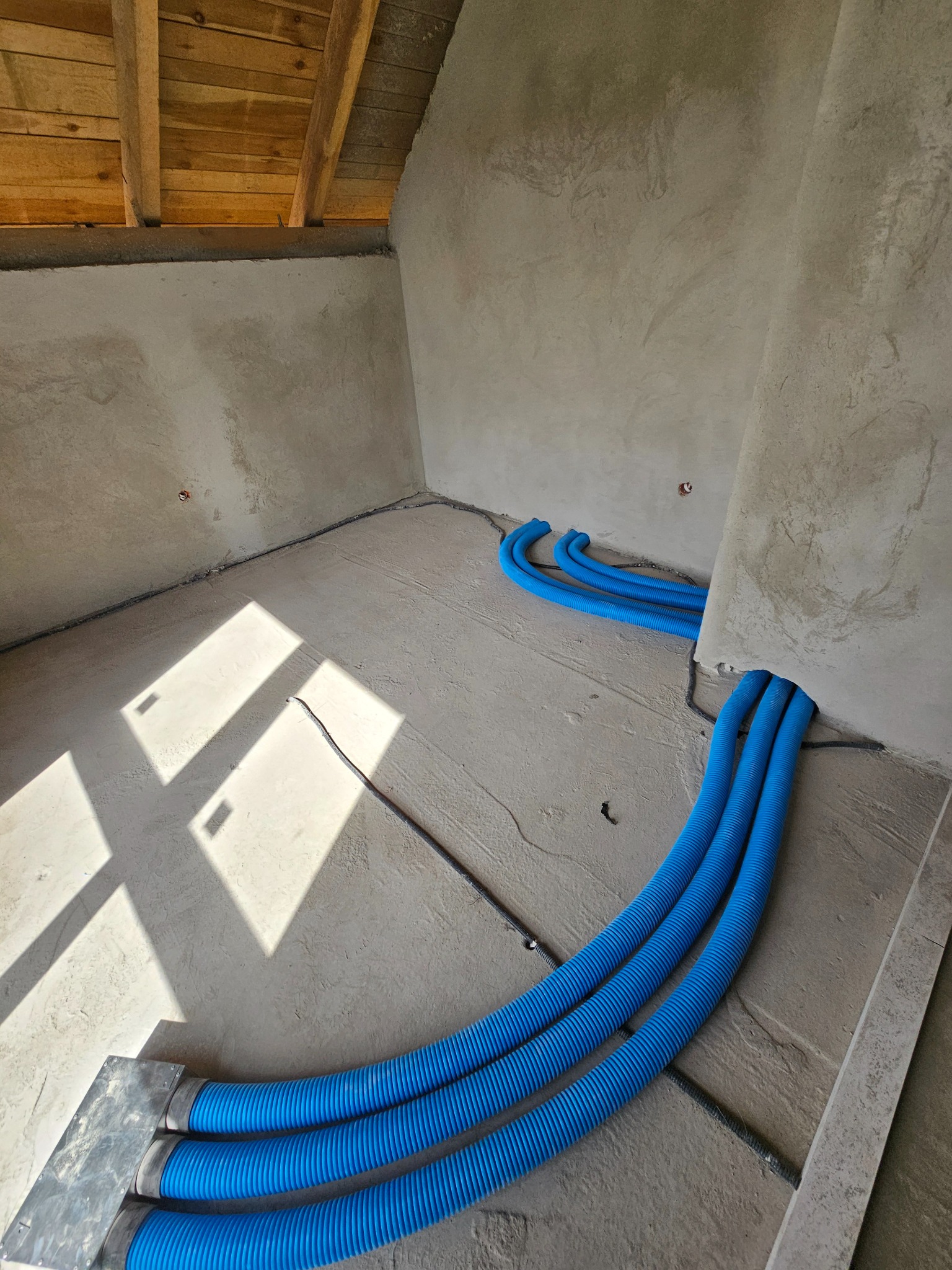 Niebieskie rury wentylacyjne ułożone na surowej posadzce w jasnym pomieszczeniu z surowymi ścianami i drewnianym stropem. Instalacja w trakcie realizacji.