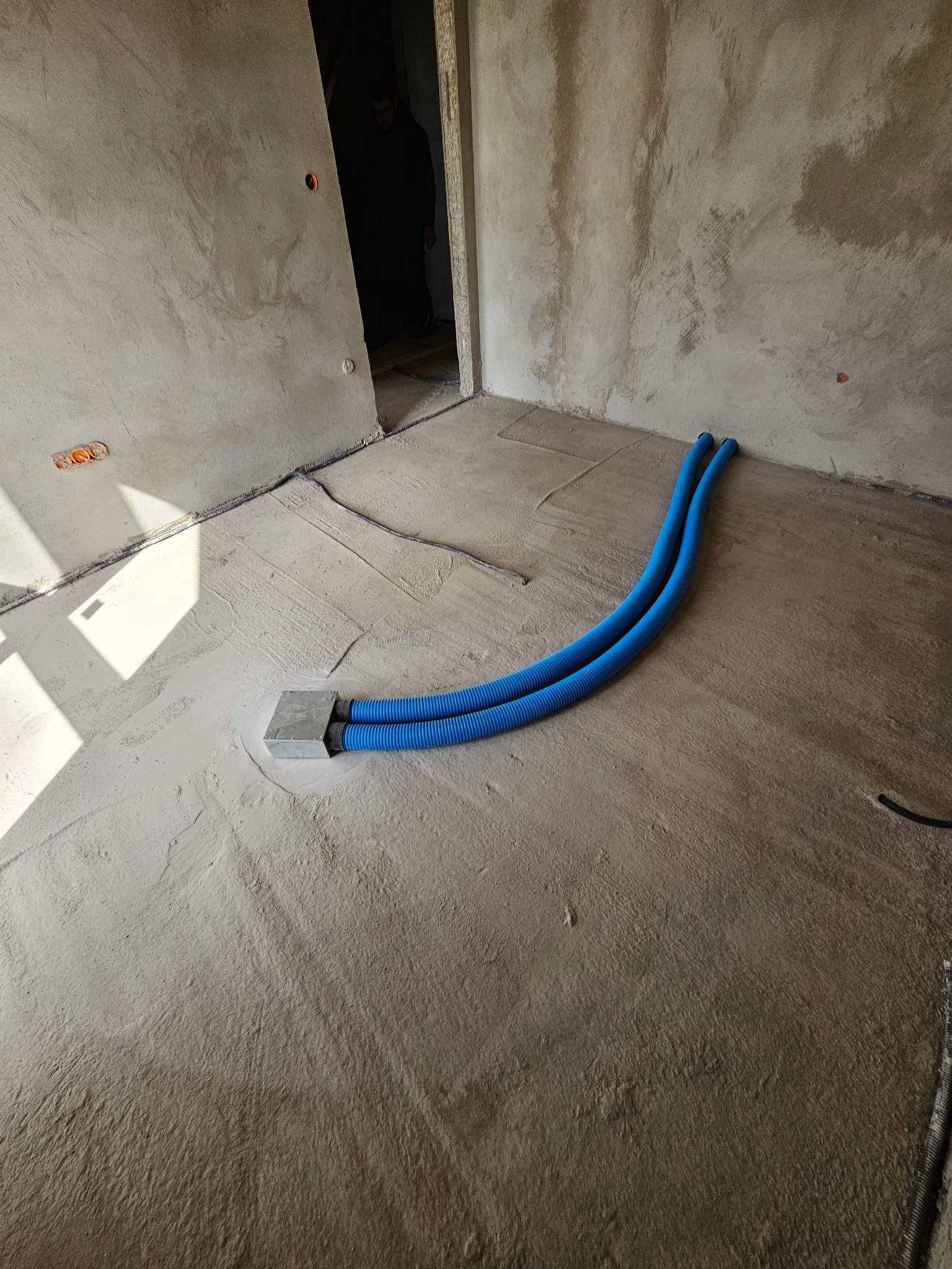 Niebieskie rury wentylacyjne wystają z podłogi w surowym pomieszczeniu. Widoczne ślady po wylewce i instalacja elektryczna w ścianie.