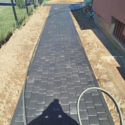 Piotr Romanowski - Ścieżka z grafitowej kostki brukowej, obrzeża z ziemi, cień osoby widoczny na kostce, fragment budynku z ceglaną podmurówką w tle.