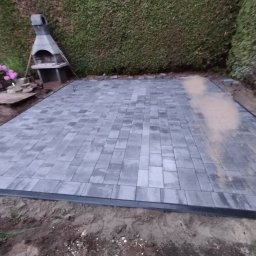 Piotr Romanowski - Ułożona, szara kostka brukowa na przygotowanym terenie. W tle zielony żywopłot i grill ogrodowy. Częściowo widoczne różowe kwiaty i ziemia.