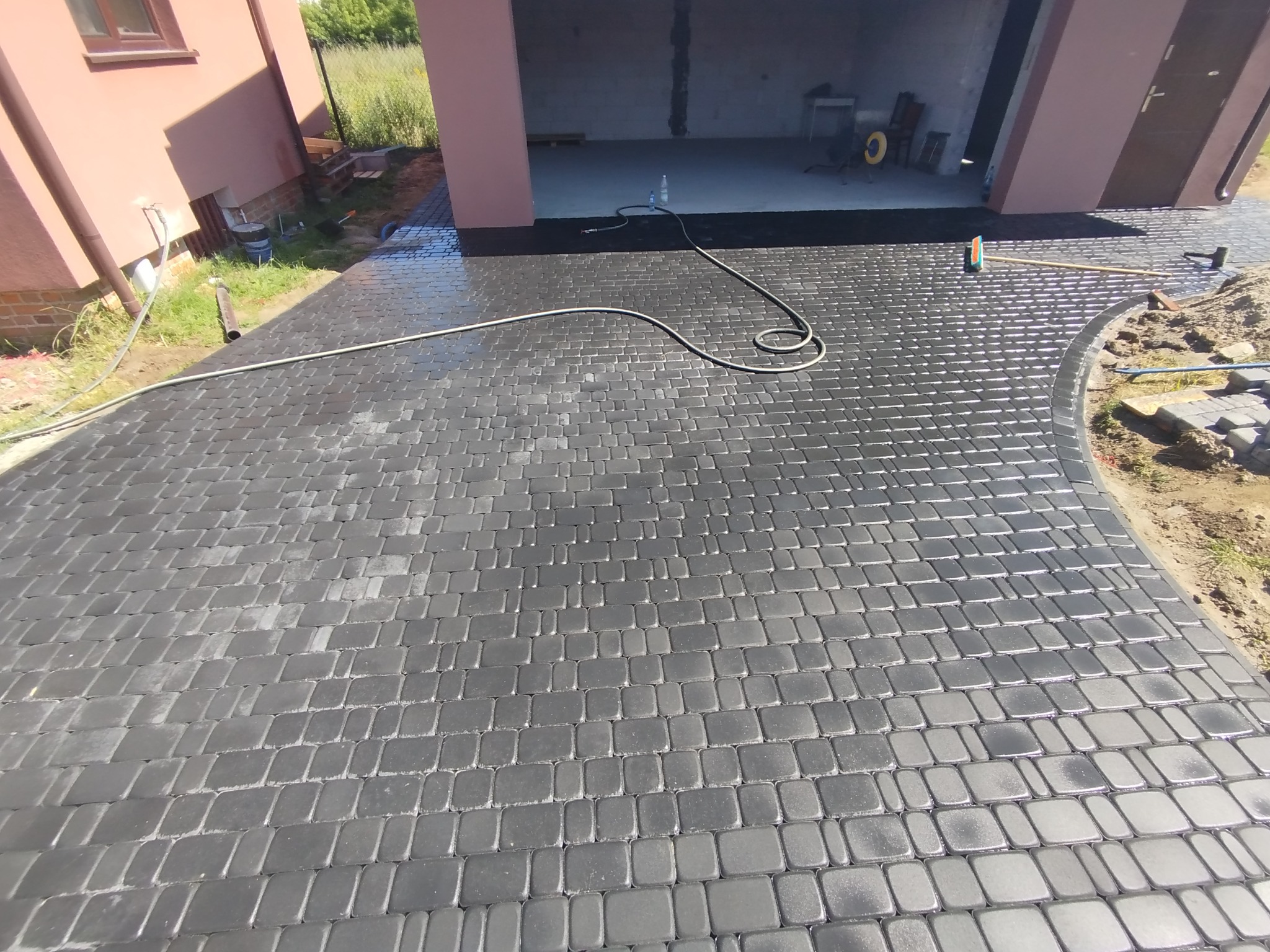 Świeżo ułożona, mokra kostka brukowa w odcieniach szarości prowadząca do garażu. Widoczne narzędzia brukarskie i wąż ogrodowy. Częściowo ułożony obrzeże.