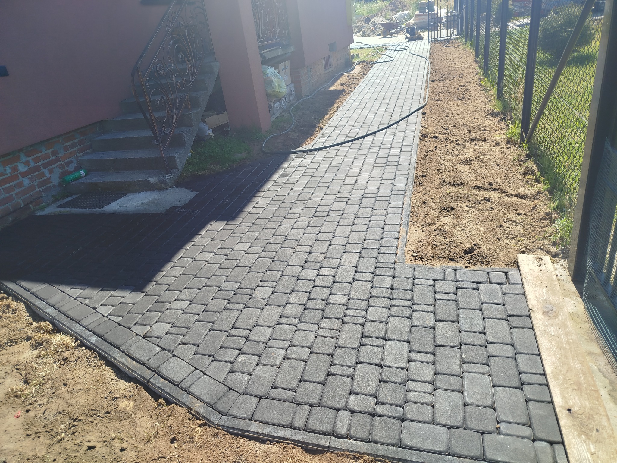 Ułożona, szara kostka brukowa tworzy ścieżkę od schodów domu do furtki. Widoczne obrzeża i ziemia po bokach. W tle narzędzia i ogrodzenie z siatki.