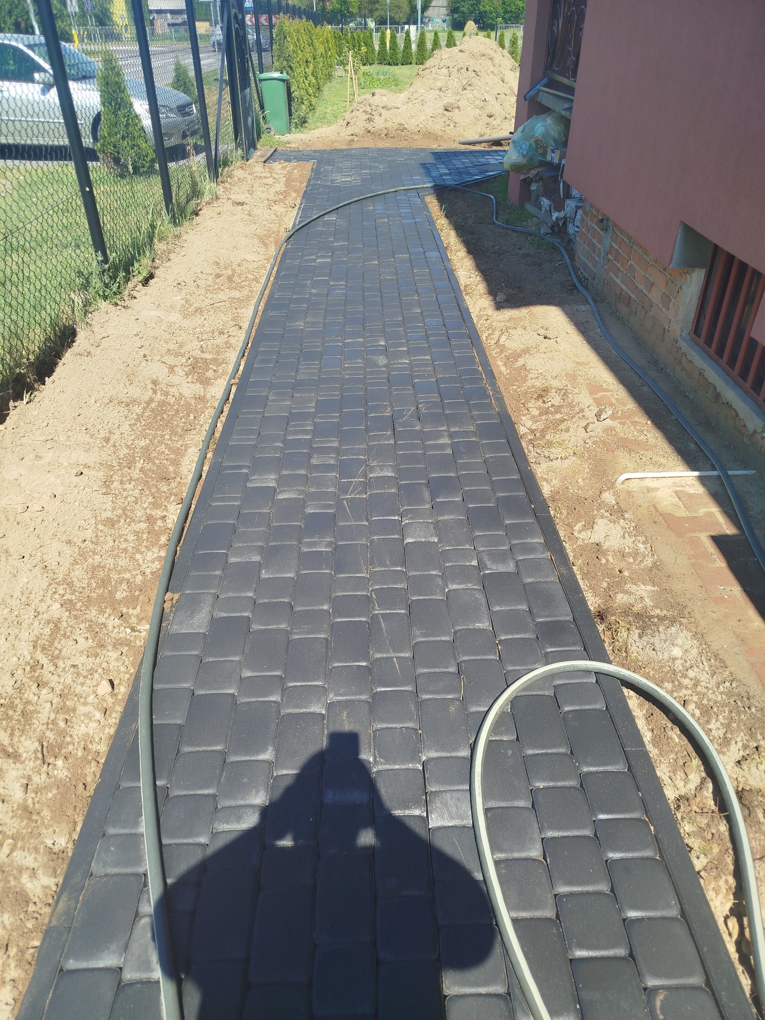 Ścieżka z grafitowej kostki brukowej, obrzeża z ziemi, cień osoby widoczny na kostce, fragment budynku z ceglaną podmurówką w tle.