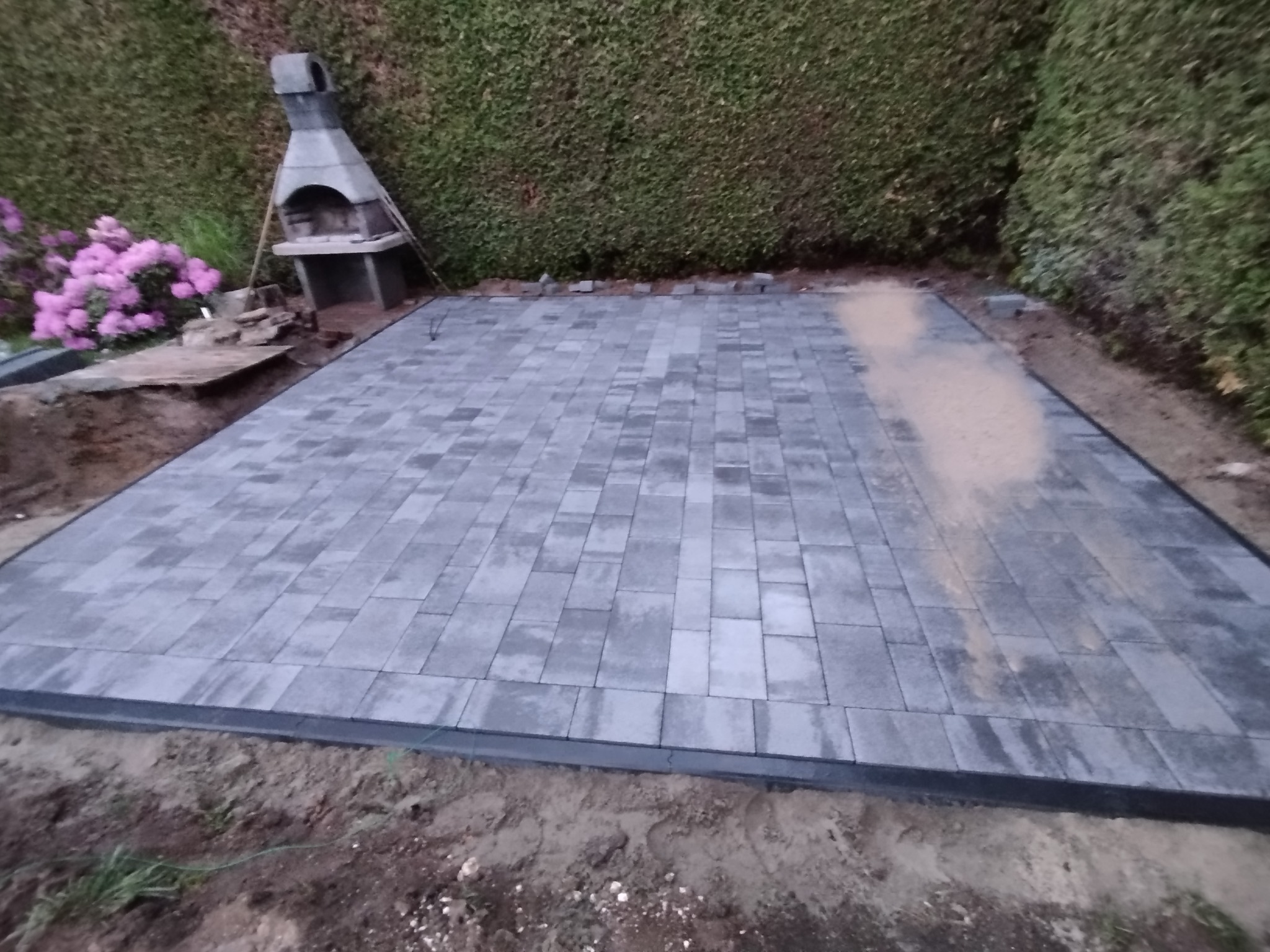 Ułożona, szara kostka brukowa na przygotowanym terenie. W tle zielony żywopłot i grill ogrodowy. Częściowo widoczne różowe kwiaty i ziemia.