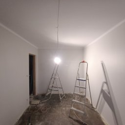 Renovacion Remonty - Wnętrze pokoju po malowaniu ścian na biało, widoczne dwie drabiny malarskie, podłoga zabezpieczona folią malarską, listwy przypodłogowe pomalowane.
