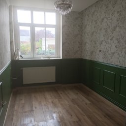 Renovacion Remonty - Pokój po remoncie: drewnopodobne panele, ściany z tapetą w roślinny wzór, lamperia w kolorze butelkowej zieleni. Kot na parapecie obserwuje otoczenie.