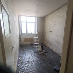 Renovacion Remonty - Pokój w trakcie szpachlowania ścian: zabezpieczona podłoga, drabina, worki z gładzią. Widoczne nierówności na ścianach przed ostatecznym wygładzeniem.