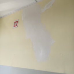 Renovacion Remonty - Żółta ściana z nałożoną warstwą białej gładzi, widoczny fragment sufitu i godło Polski w ramce. Etap prac przygotowawczych do malowania.