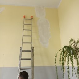 Renovacion Remonty - Żółta ściana z szarym dołem, częściowo wyszpachlowana, drabina, mężczyzna w okularach. W rogu zielona roślina doniczkowa.