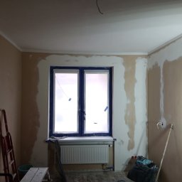 Renovacion Remonty - Pokój w trakcie szpachlowania ścian, okno zabezpieczone folią, widoczne narzędzia i drabina. Ściany częściowo pokryte gładzią, sufit pomalowany na biało.