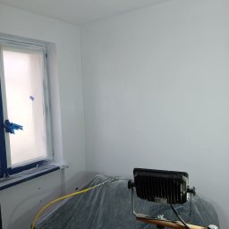 Renovacion Remonty - Świeżo pomalowane na biało ściany w pokoju w Kobylance, okno zabezpieczone folią malarską, lampa oświetla wnętrze. Widoczne ślady po szpachlowaniu.
