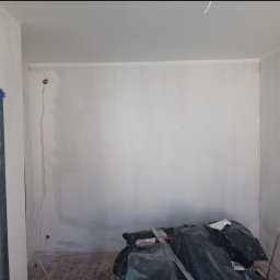 Renovacion Remonty - Pokój w trakcie szpachlowania ścian, widoczne narzędzia i worki z odpadami. Otwór drzwiowy zabezpieczony folią malarską. Ściany przygotowane do dalszych prac.