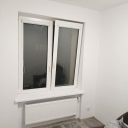 Renovacion Remonty - Białe, plastikowe okno uchylone w jasnym pokoju z grzejnikiem pod parapetem. Ściany pomalowane na biało, widoczny fragment podłogi i sofy.