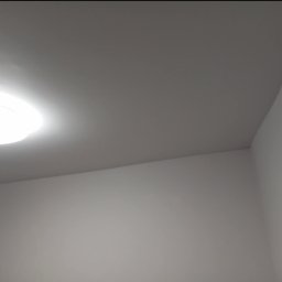 Renovacion Remonty - Gładka, pomalowana na szaro ściana w rogu pomieszczenia, oświetlona okrągłą lampą sufitową. Minimalistyczne wykończenie wnętrza.