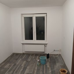 Renovacion Remonty - Pusty pokój po odświeżeniu: białe ściany, szara podłoga imitująca drewno, okno z grzejnikiem. Widoczne akcesoria do sprzątania: wiadro i miska.