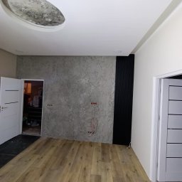 Renovacion Remonty - Pokój po wykończeniu: drewnopodobna podłoga, ściana z szarą fakturą, czarne panele dekoracyjne, białe drzwi i sufit z okrągłym wgłębieniem. Widoczne kable elektryczne.