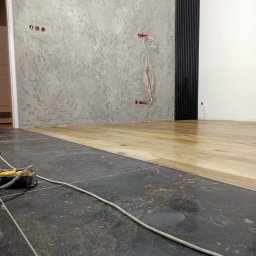Renovacion Remonty - Wnętrze w trakcie prac: połączenie szarej ściany z dekoracyjną fakturą, czarnych paneli ściennych i jasnej drewnianej podłogi. Widoczne kable elektryczne i narzędzia.