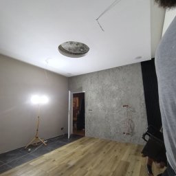 Renovacion Remonty - Wykończone wnętrze z drewnianą podłogą, szarą ścianą z fakturą i białym sufitem z otworem na oświetlenie. Widoczna lampa na statywie i fragment osoby.
