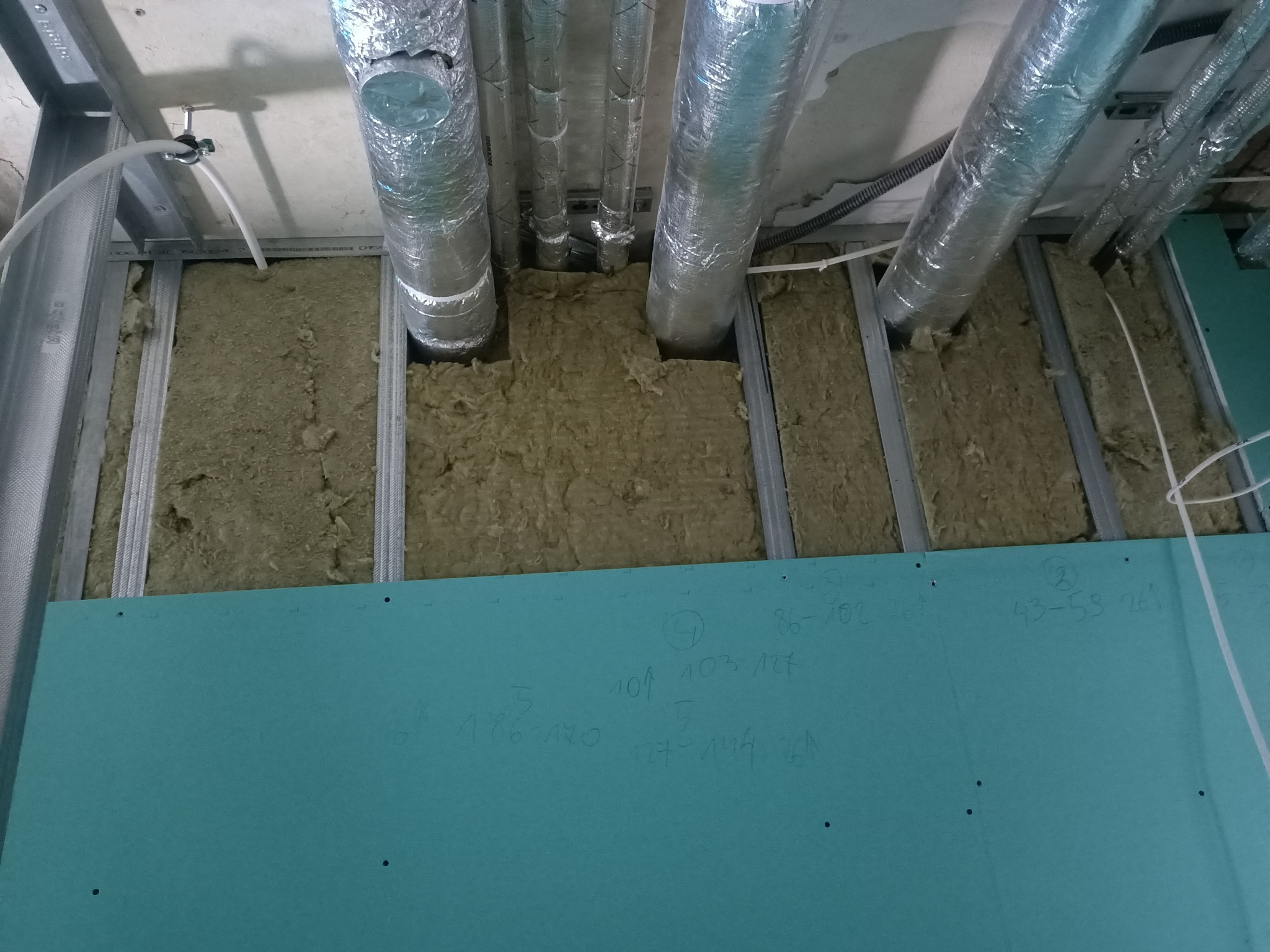Montaż instalacji: wełna mineralna w metalowym stelażu, rury wentylacyjne w izolacji, zielona płyta gipsowo-kartonowa z odręcznymi notatkami.