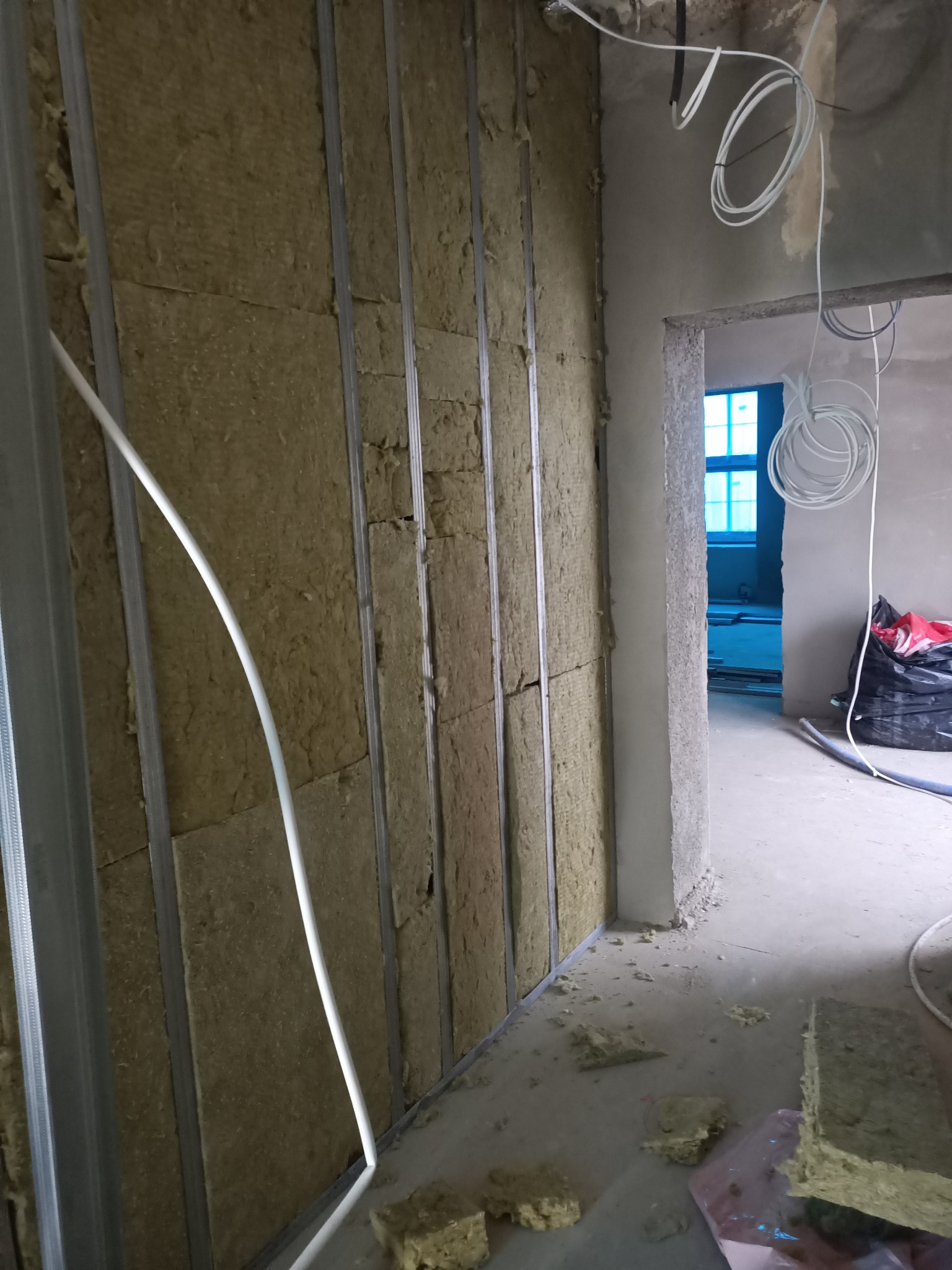 Ocieplanie ścian wełną mineralną, widoczne metalowe profile konstrukcyjne i fragment instalacji elektrycznej. W tle widoczne pomieszczenie w trakcie prac wykończeniowych.