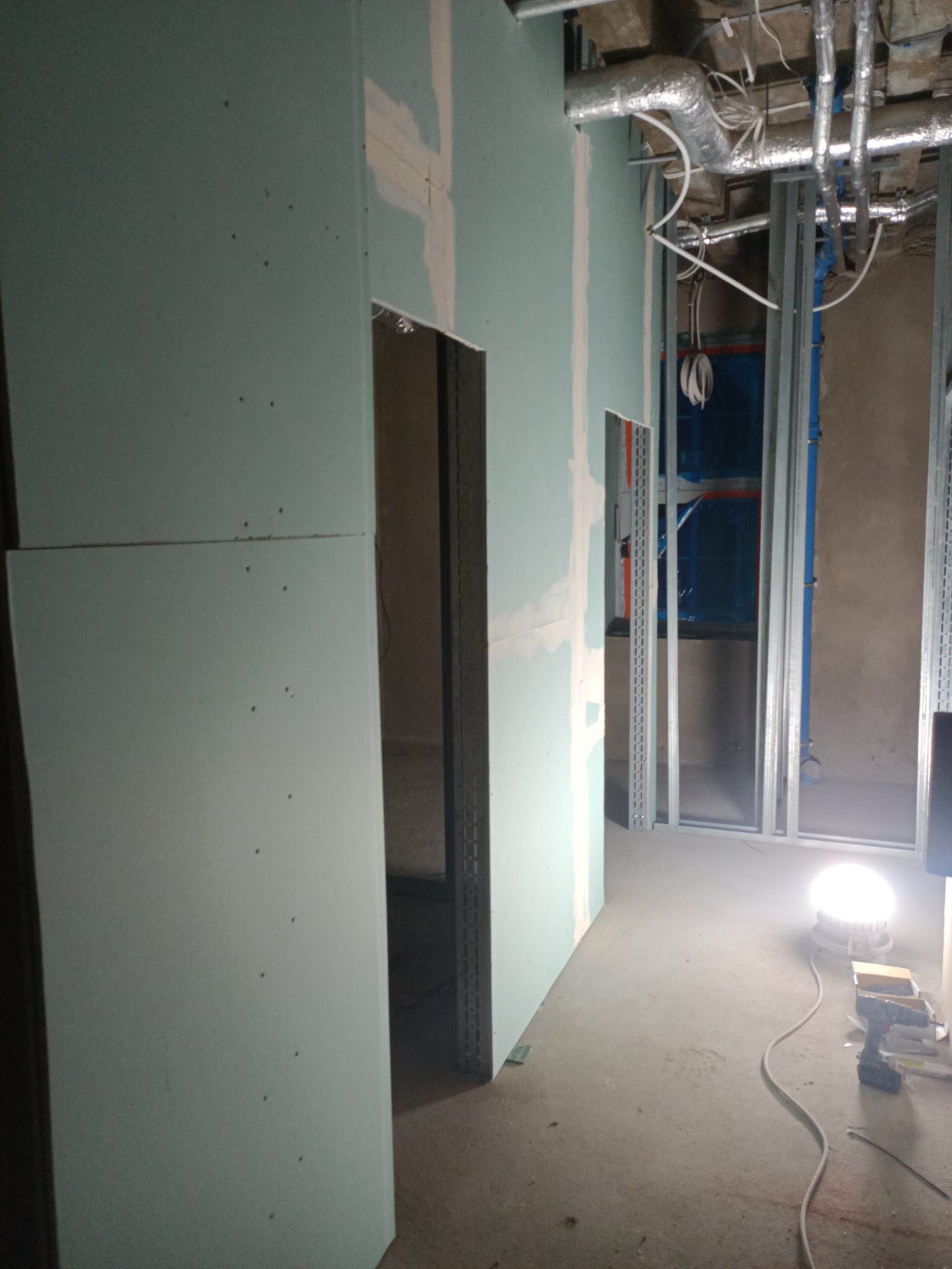 Montaż płyt gipsowo-kartonowych na metalowym stelażu, widoczne instalacje pod sufitem, narzędzia na podłodze. Prace wykończeniowe w surowym wnętrzu.