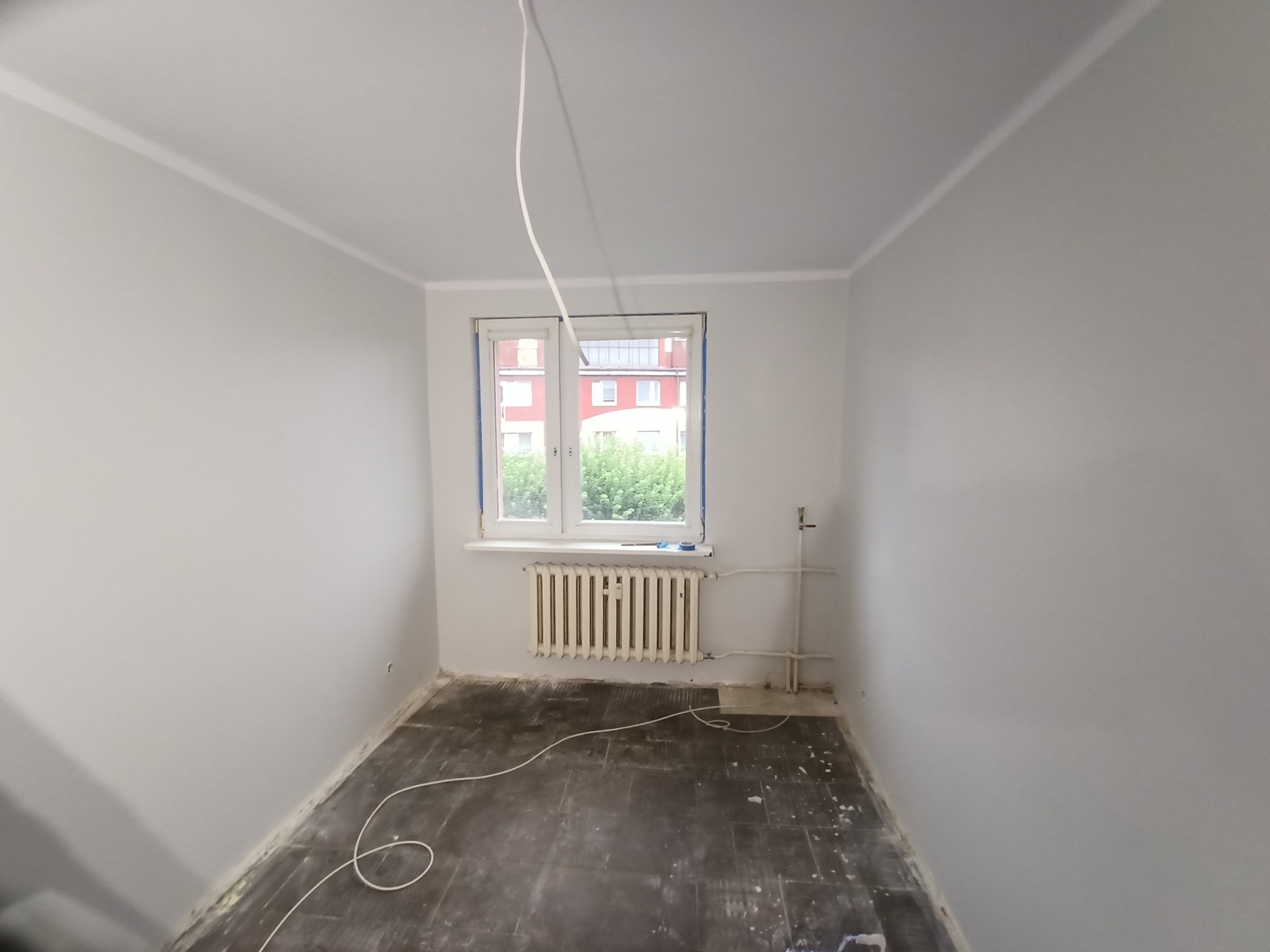 Pokój po malowaniu na szaro, z białymi listwami przysufitowymi, grzejnikiem pod oknem i wystającymi kablami. Przygotowanie do dalszych prac wykończeniowych.