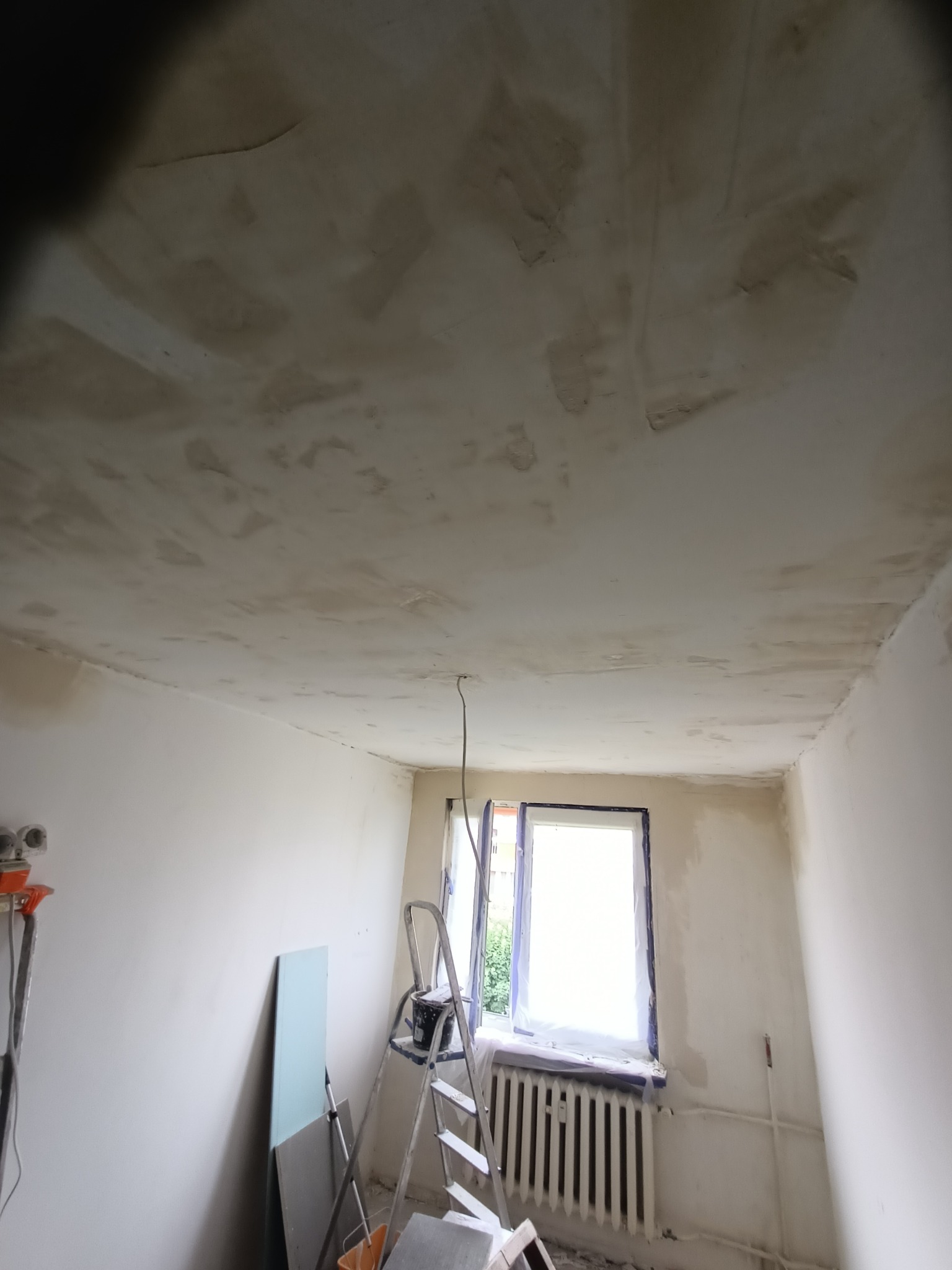 Pokój w trakcie prac wykończeniowych: sufit po szpachlowaniu, ściany przygotowane do malowania. Drabina, płyty gipsowe i narzędzia w pomieszczeniu.