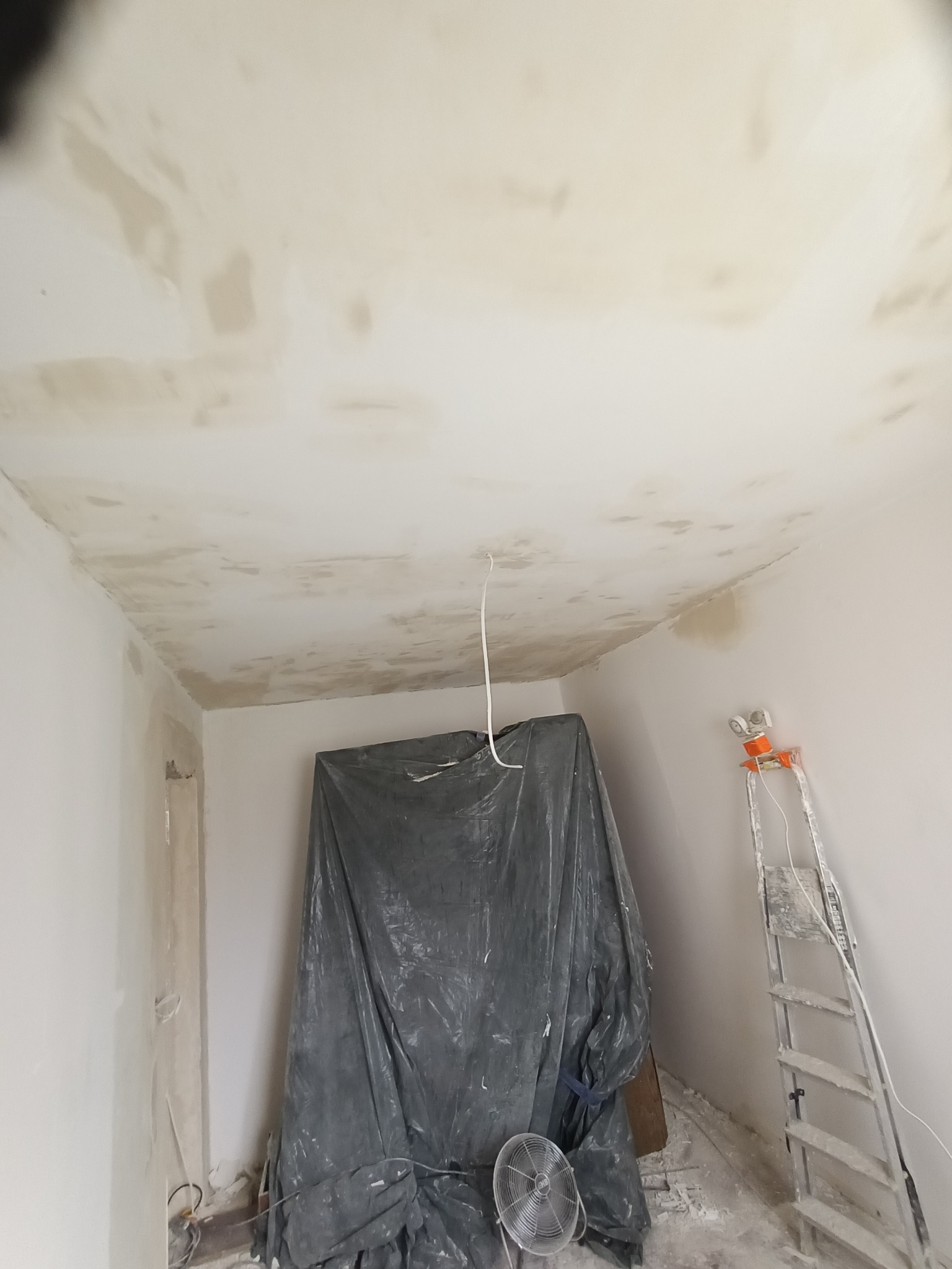 Przygotowanie sufitu do prac wykończeniowych. Widoczna folia ochronna, drabina i wentylator. Sufit po szpachlowaniu, gotowy do dalszych etapów.