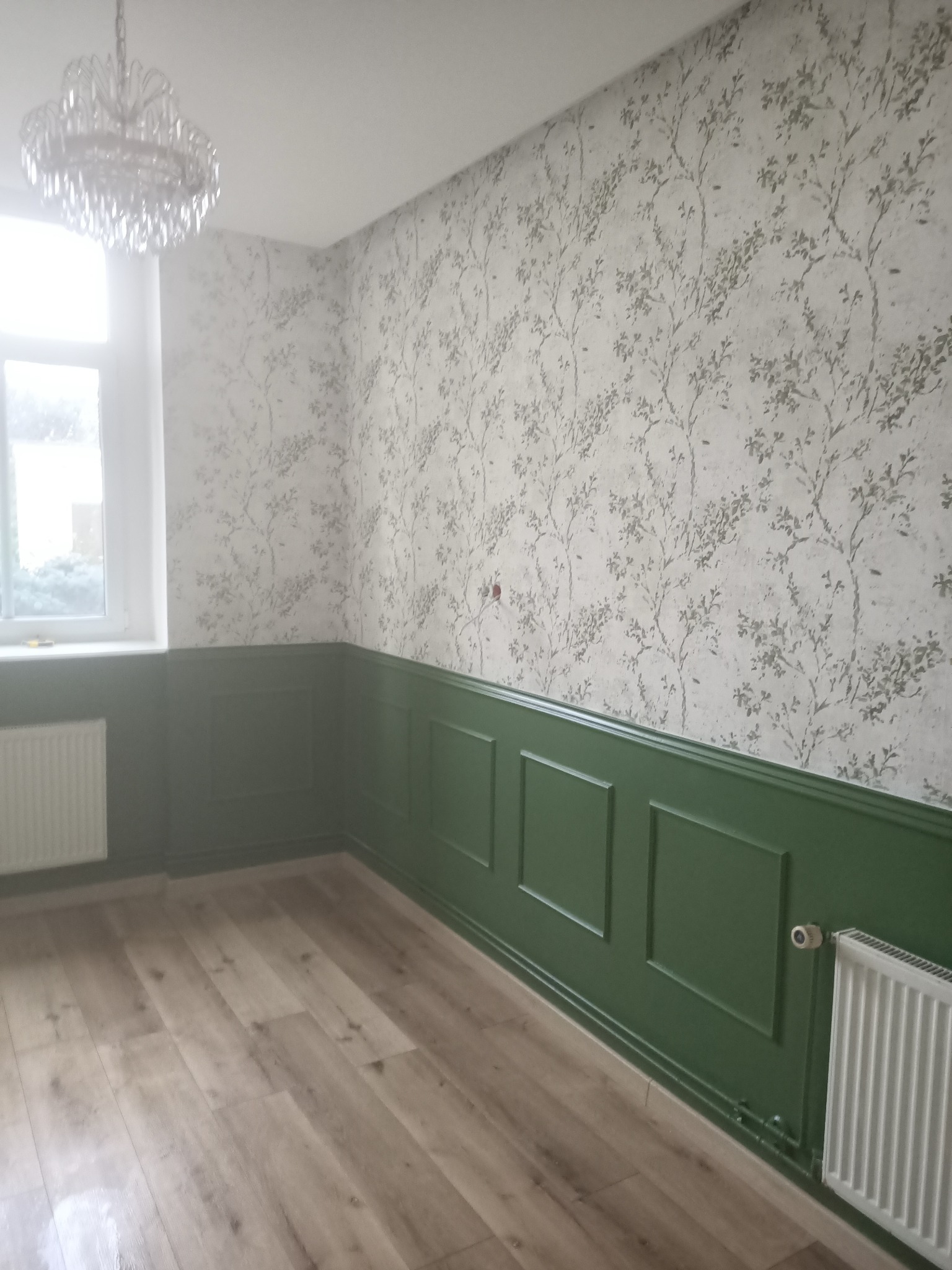 Pokój po remoncie: panele podłogowe, zielona lamperia, ściany w tapetę z motywem roślinnym. Kryształowy żyrandol i biały grzejnik.