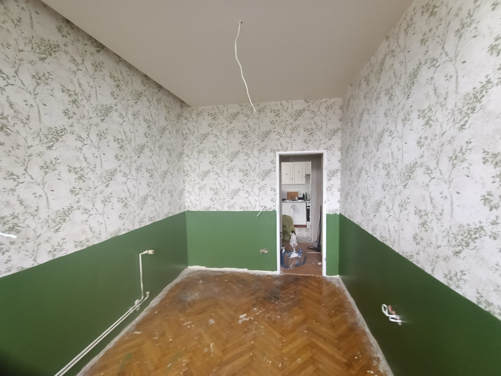 Pokój po częściowym remoncie: dolna część ścian pomalowana na zielono, górna w tapetę w delikatny wzór. Parkiet wymaga odświeżenia. Widoczne kable.