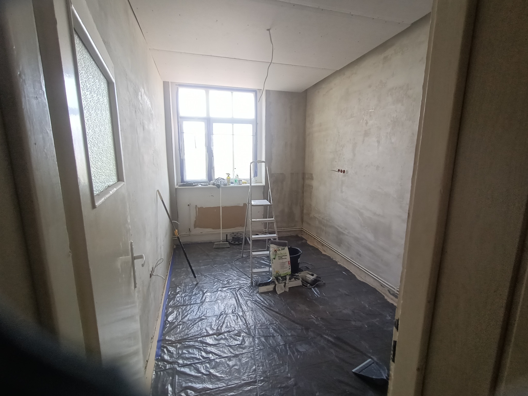 Pokój w trakcie szpachlowania ścian: zabezpieczona podłoga, drabina, worki z gładzią. Widoczne nierówności na ścianach przed ostatecznym wygładzeniem.