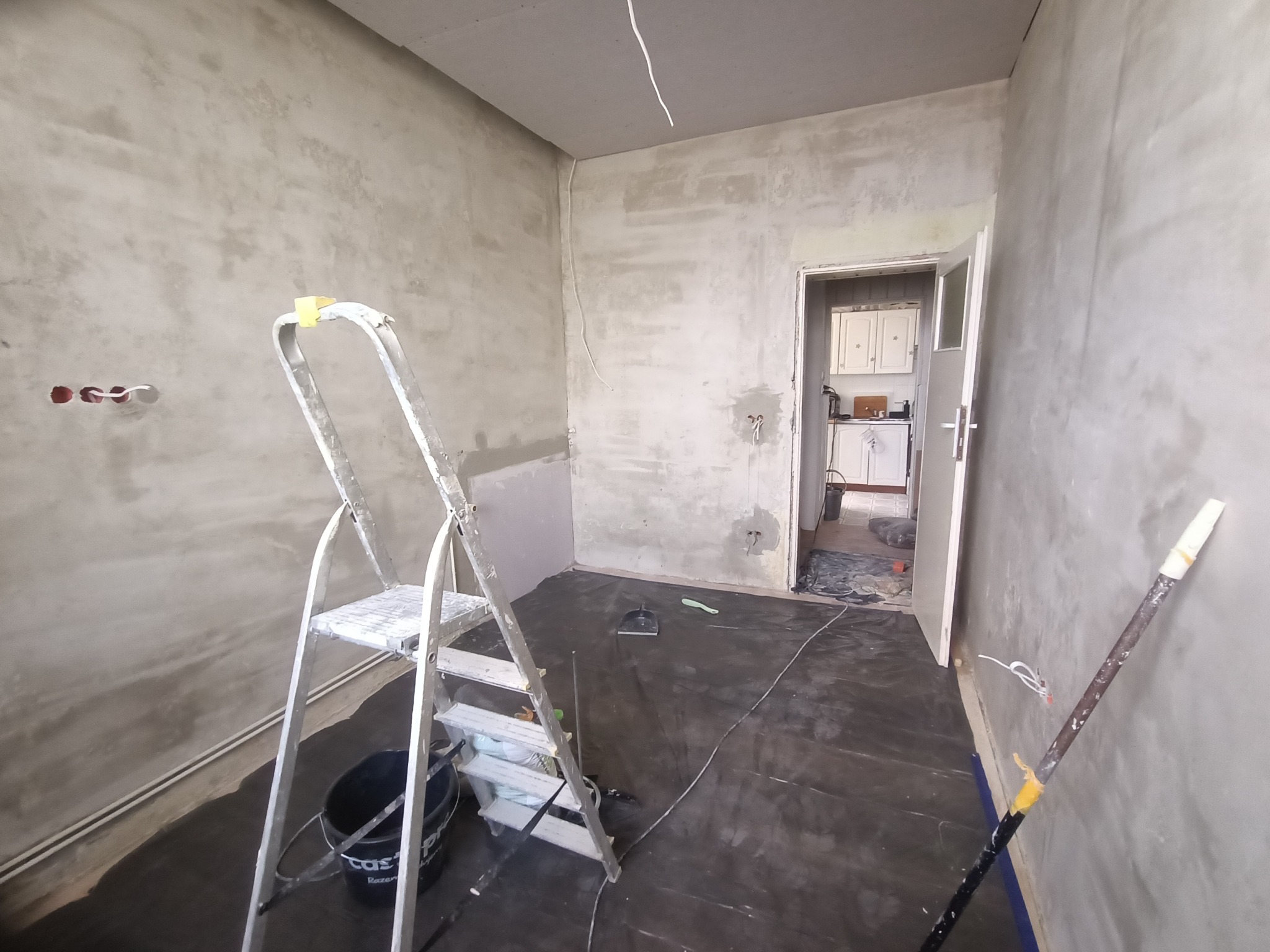 Pokój w trakcie szpachlowania ścian, widoczna drabina, wiadro i zakryta podłoga. W tle widoczny fragment kuchni. Ściany pokryte szarą masą szpachlową.