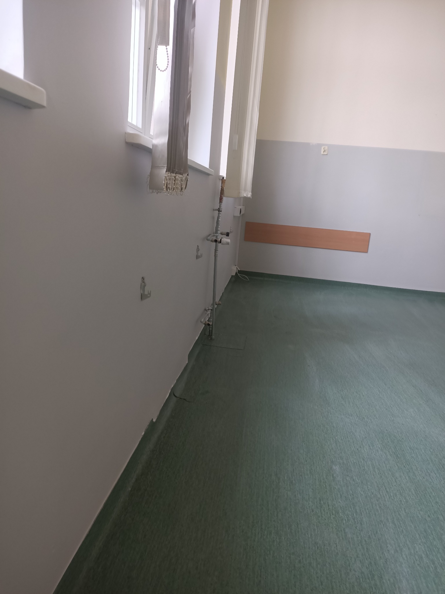 Pomalowane na szaro ściany i zielona podłoga w pustym pomieszczeniu. Widoczne rury przy ścianie, okno z żaluzjami i listwa przypodłogowa.