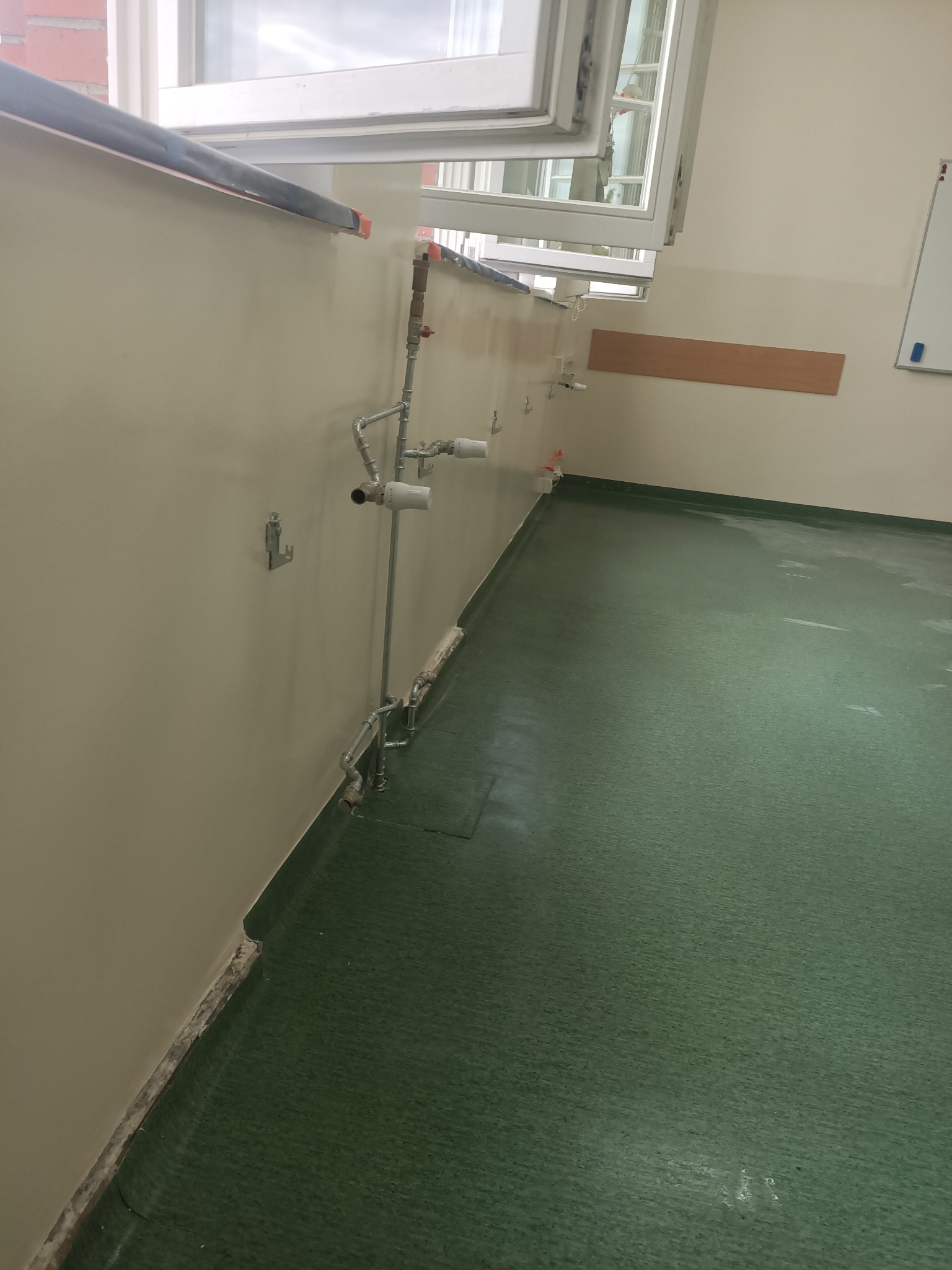 Wnętrze w trakcie prac: ściana z instalacją wodną, zielona podłoga, okno. Widoczne ślady po demontażu. Przygotowanie do dalszych prac wykończeniowych, detale instalacji.