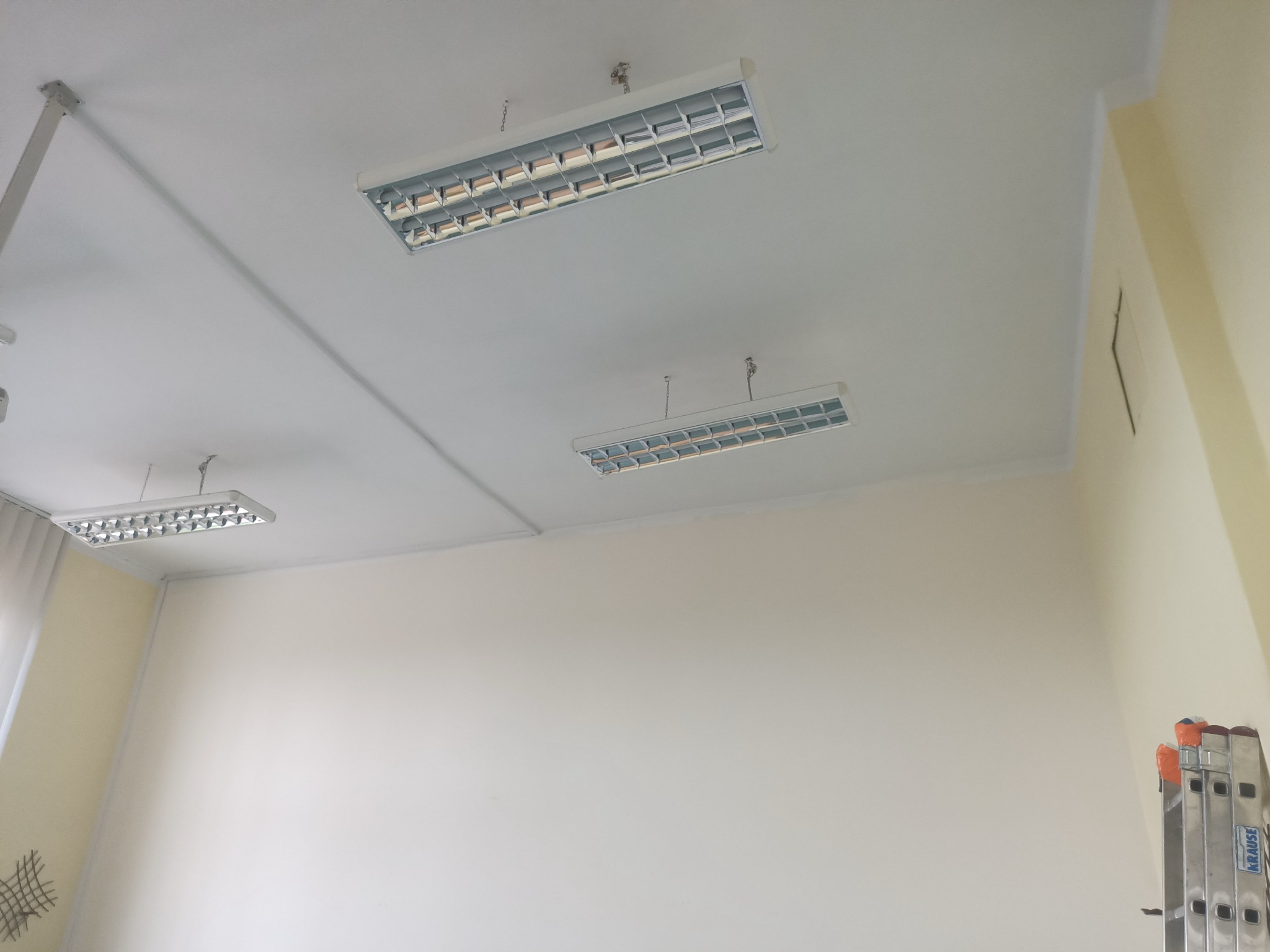 Białe, świeżo pomalowane wnętrze z trzema lampami sufitowymi i rozstawioną drabiną w rogu. Widoczny fragment ściany w odcieniu kremowym.