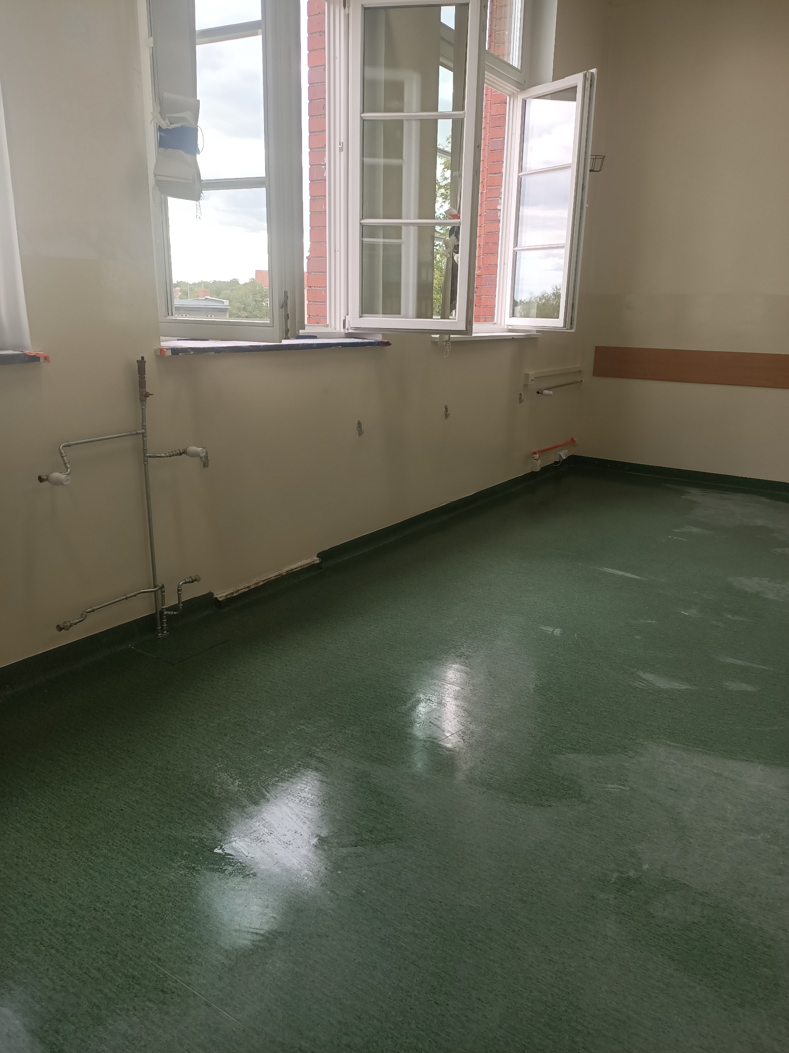 Pokój po remoncie: świeżo pomalowane ściany w odcieniach beżu, zielona podłoga, białe okna z widokiem na zieleń. Widoczne rury instalacji wodnej.