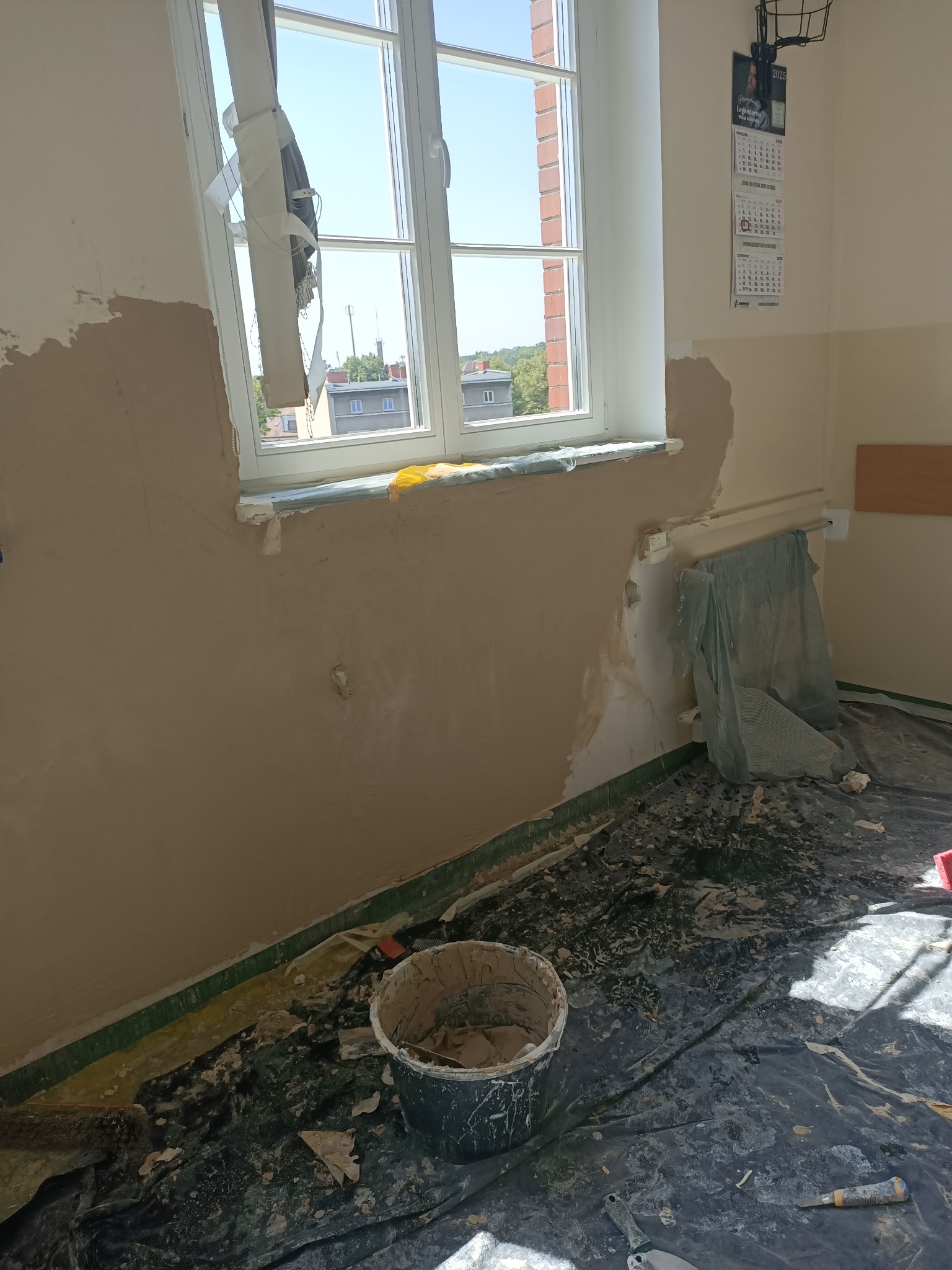Pokój w trakcie szpachlowania ścian, widoczne narzędzia, wiadro z gładzią i zabezpieczona folią podłoga. Częściowo zaszpachlowana ściana przy oknie.