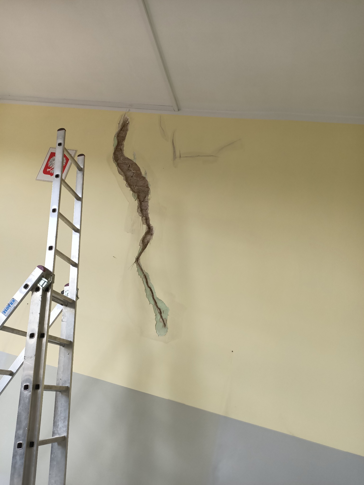 Ściana z pęknięciem w trakcie naprawy, widoczna drabina Krause i częściowo usunięta farba. Dół ściany pomalowany na szaro, góra na żółto. Sufit biały.