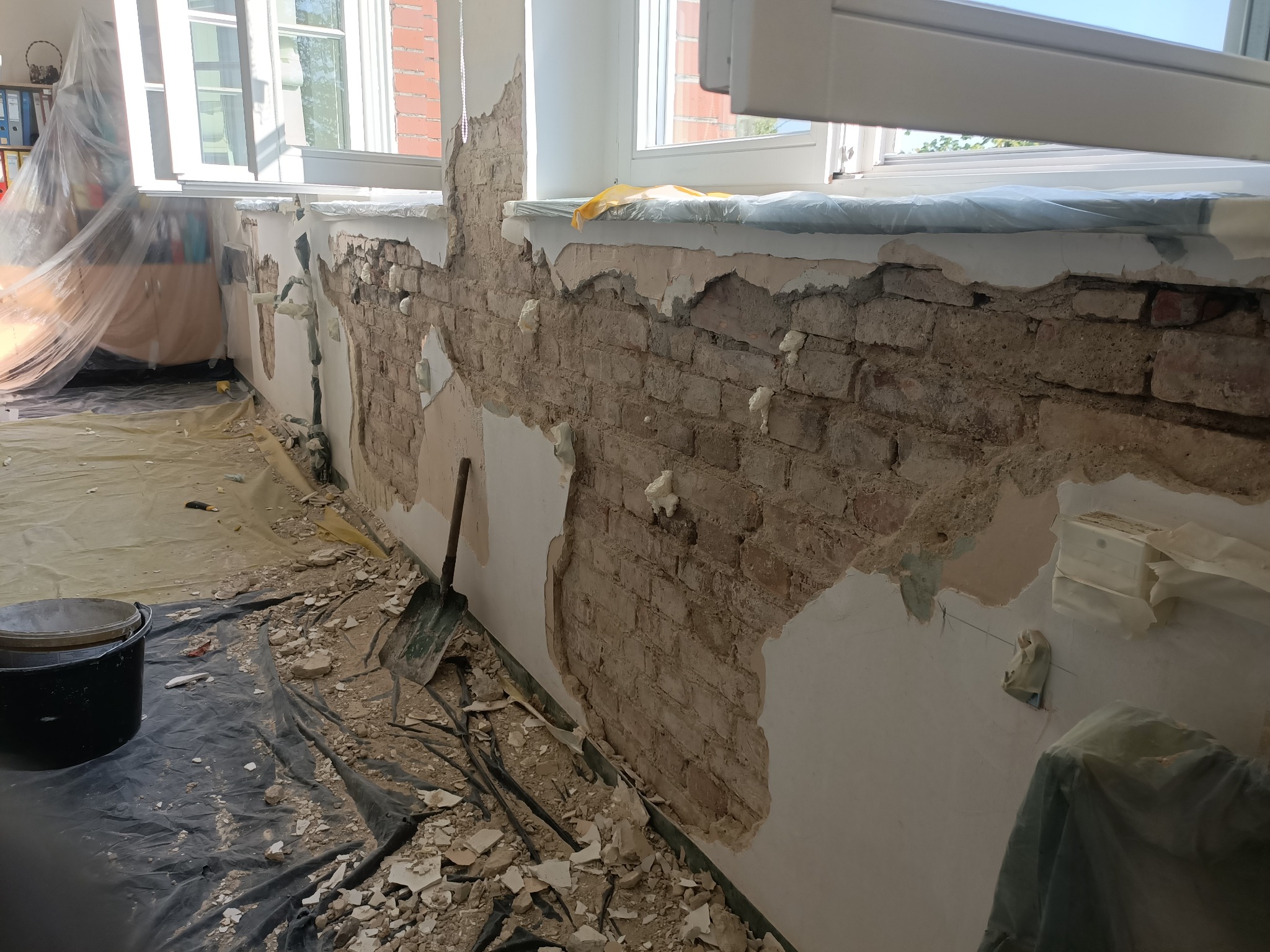 Odsłonięta ceglana ściana w trakcie renowacji, z fragmentami starego tynku i narzędziami na podłodze. Widoczne okna i zabezpieczone powierzchnie.